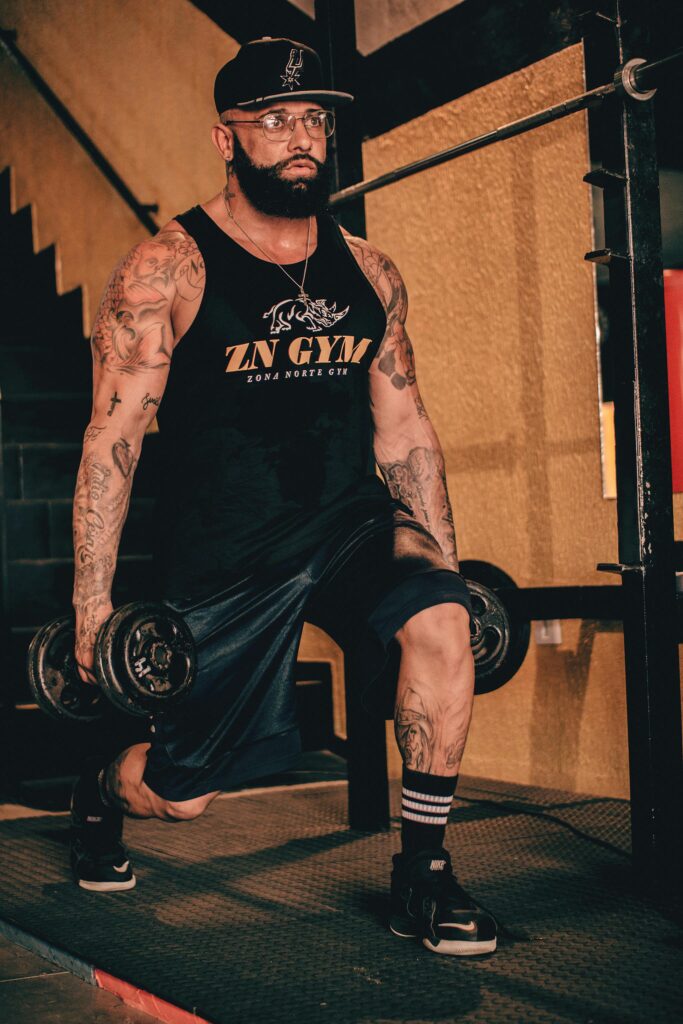 Tattooed man in black tank top lunging with dumbbells in gym setting.