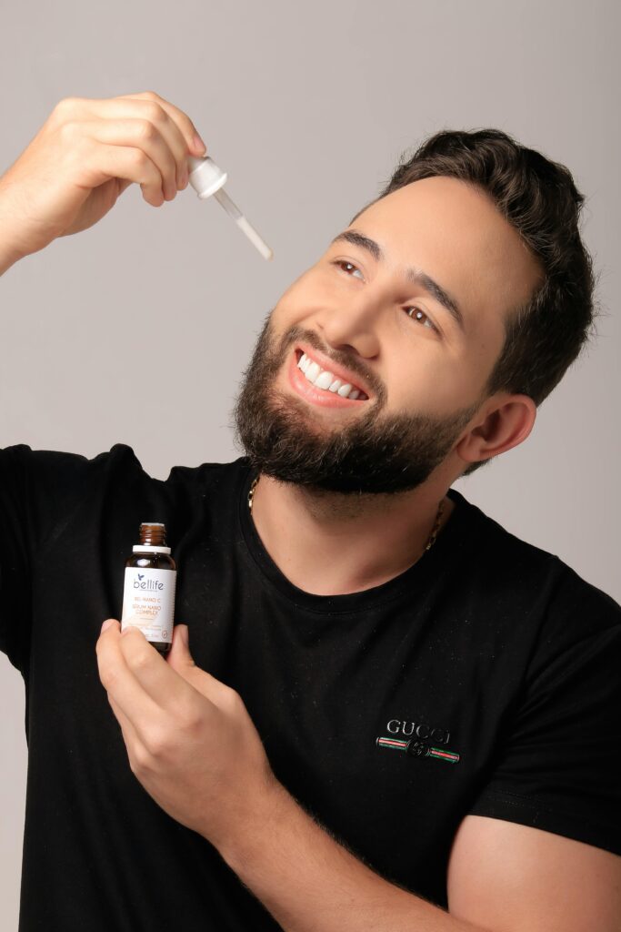 Smiling man applying beard oil with a dropper against a grey background.
