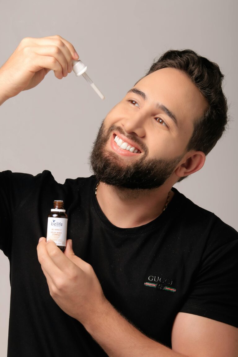 Smiling man applying beard oil with a dropper against a grey background.